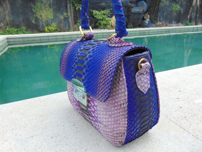 May include: A small, purple and blue handbag made of snakeskin. The bag has a top handle and a flap closure.