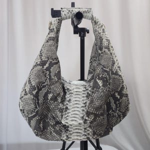 Peut inclure: Un sac à main de style hobo avec un motif en peau de serpent gris et blanc. Le sac présente une silhouette souple et non structurée avec une poignée intégrée unique et une texture d'écailles verticale centrale sur le panneau avant.
