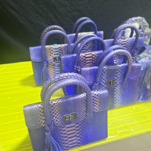 Peut inclure: Une collection de sacs à main structurés présentant un motif texturé en forme d'écailles dans des tons de bleu et de violet. Chaque sac à main comprend une poignée supérieure, une fermeture à tourniquet rectangulaire argentée et un pompon.