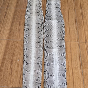 May include: Two pieces of snakeskin leather, featuring a pattern of gray, black, and white scales. The leather is laid out on a wooden surface. The skins have a central white stripe.