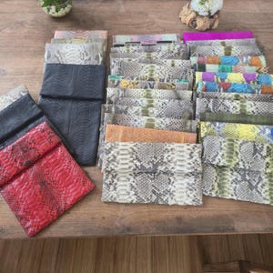 Könnte beinhalten: Eine Sammlung rechteckiger Clutch-Taschen mit verschiedenen Schlangenhautmustern in Farben wie Rot, Schwarz, Gelb, Blau, Grün und natürlichen Grautönen. Die Taschen sind in mehreren Stapeln angeordnet und zeigen vielfältige Texturen und Schuppen.
