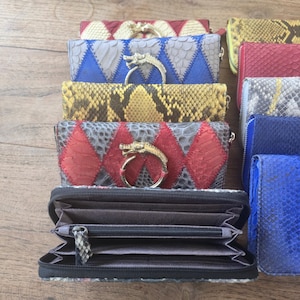 May include: A collection of rectangular wallets featuring textured snakeskin patterns in various colors including red, blue, yellow, and gray. Several wallets are adorned with a metallic dragon-shaped ring pull. One wallet is open, revealing a dark gray interior with multiple card slots and a central zippered compartment.