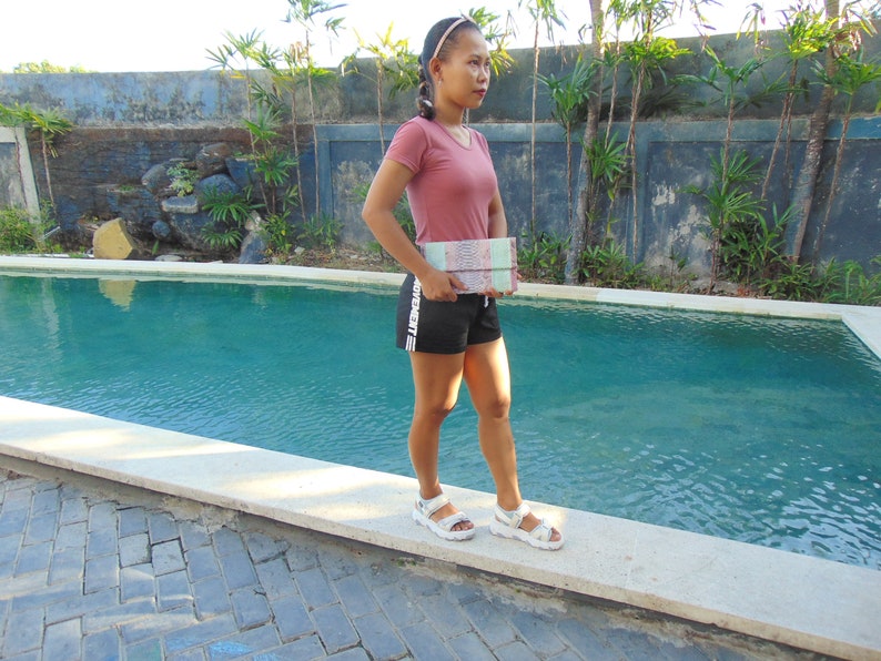 Pu&ograve; includere: Una donna con una camicia rosa e pantaloncini neri sta in piedi vicino a una piscina. Tiene una pochette a mano con un motivo.