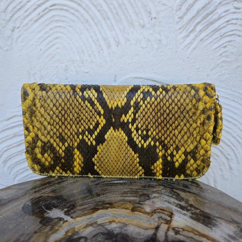 Puede incluir: Una cartera de piel de serpiente amarilla y negra con cierre de cremallera.