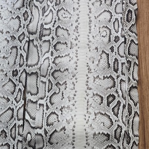 May include: Close-up of a snakeskin pattern in shades of white, grey, and black. The design features a repeating pattern of irregular shapes, creating a textured appearance. The skin is likely used for fashion or craft projects.