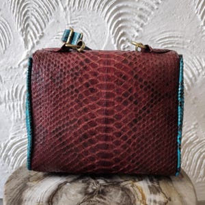 May include: A burgundy leather handbag with a turquoise trim and a snakeskin pattern. The bag has a shoulder strap and a top flap closure.