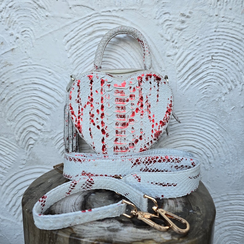 Pu&ograve; includere: Una borsa a forma di cuore con un motivo a pelle di serpente bianco e rosso. La borsa ha un manico superiore, una tracolla staccabile e finiture color oro. La borsa misura circa 20 cm di larghezza e 18 cm di altezza.