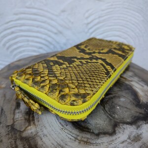 Puede incluir: Una cartera de piel de serpiente amarilla con una cremallera amarilla brillante. La cartera est&aacute; hecha de cuero genuino y tiene un patr&oacute;n &uacute;nico y texturizado.