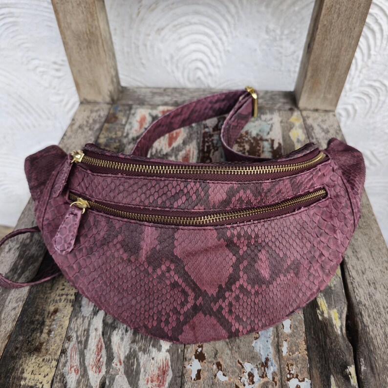 May include: A burgundy fanny pack with a snakeskin pattern. The bag has three zippered compartments with gold-colored zippers and a matching adjustable strap. The fanny pack is made of a textured material and is sitting on a wooden chair.