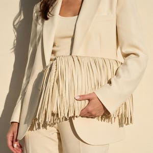 May include: A cream-colored clutch bag featuring long, layered fringe detailing across the front. The accessory is paired with a matching cream-colored blazer and top, creating a monochromatic look with a textured, layered fringe finish.