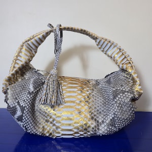 May include: A handbag with a snakeskin pattern in shades of gray, white, and gold. The bag has a curved handle and a tassel. The bag's design features a mix of textures and colors.
