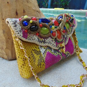 May include: A yellow, pink and white snakeskin clutch bag with a gold chain strap. The bag is decorated with a large, colourful beaded embellishment featuring a dragonfly design.