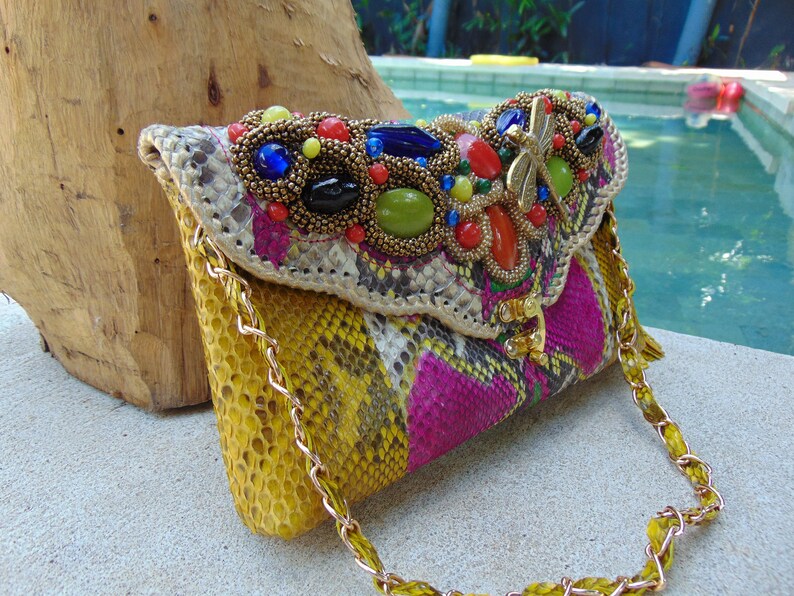 May include: A yellow, pink and white snakeskin clutch bag with a gold chain strap. The bag is decorated with a large, colourful beaded embellishment featuring a dragonfly design.
