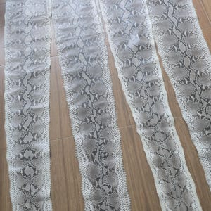 Op de afbeelding: Vier lange, smalle stukken slangenleer met een grijs en wit patroon liggen op een houten oppervlak. De huiden hebben een natuurlijk, getextureerd uiterlijk, met een schubachtig ontwerp. De huiden zijn waarschijnlijk voor knutsel- of decoratieve doeleinden.