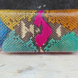 K&ouml;nnte beinhalten: Eine farbenfrohe Schlangenleder-Geldb&ouml;rse mit Rei&szlig;verschluss. Die Geldb&ouml;rse ist aus echtem Pythonleder gefertigt und hat ein geometrisches Muster in Blaut&ouml;nen, Gr&uuml;n, Gelb und Pink.