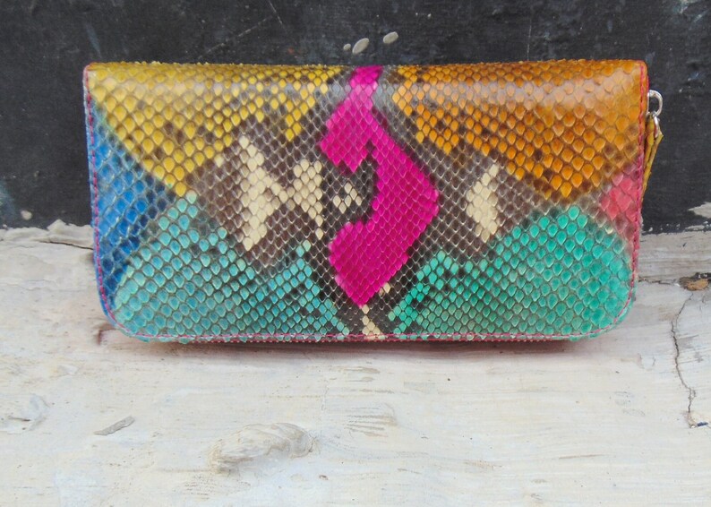 K&ouml;nnte beinhalten: Eine farbenfrohe Schlangenleder-Geldb&ouml;rse mit Rei&szlig;verschluss. Die Geldb&ouml;rse ist aus echtem Pythonleder gefertigt und hat ein geometrisches Muster in Blaut&ouml;nen, Gr&uuml;n, Gelb und Pink.