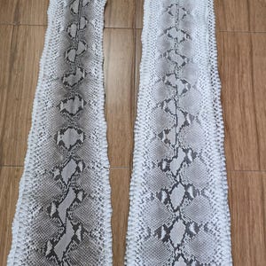 Peut inclure: Deux pièces de cuir de peau de serpent, présentant un motif d'écailles grises, marron et blanches. Le cuir est disposé sur une surface en bois, mettant en valeur la texture naturelle et les variations de couleur.