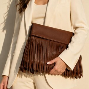 May include: A brown leather clutch with long fringe detail. The clutch is held against a cream-colored blazer and pants. The bag's fringe hangs down, creating a textured look.
