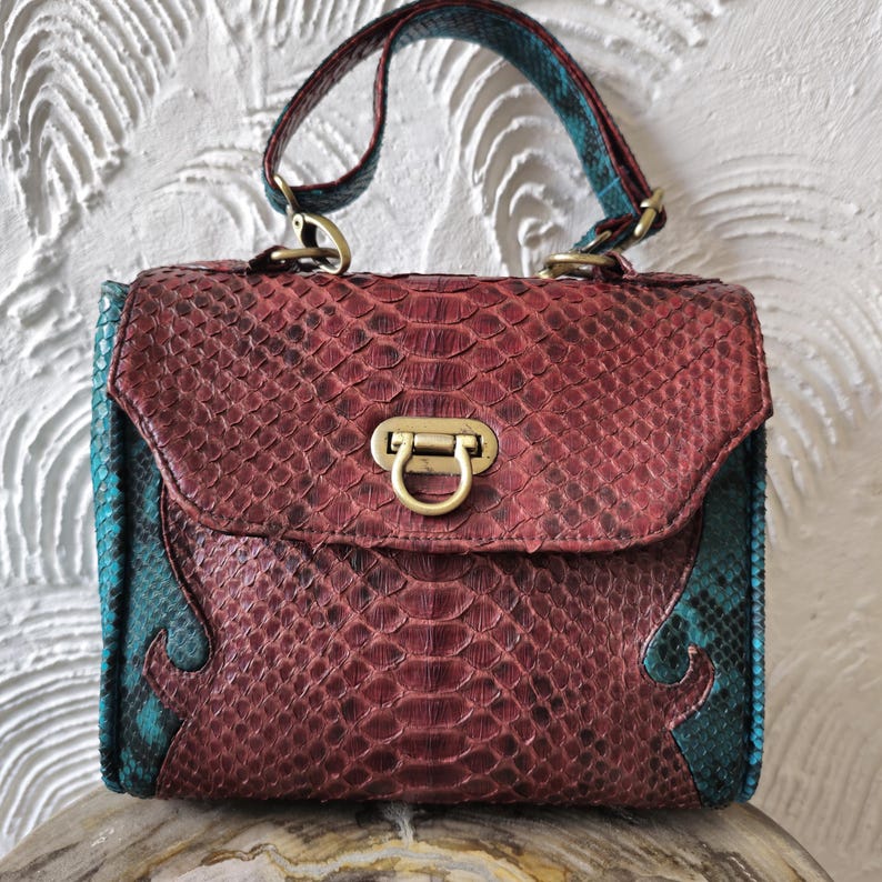 May include: A burgundy and teal snakeskin handbag with a brass clasp and a top handle.