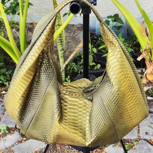 May include: A hobo-style handbag crafted from textured, metallic olive-green snakeskin leather. The bag features a single integrated shoulder strap, a slouchy crescent shape, and a zippered top closure. The scales are visible across the entire surface.