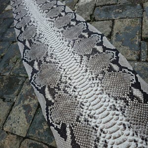 May include: A snakeskin pattern in shades of gray, black, and white. The pattern is detailed with scales and markings, creating a realistic texture. The snakeskin is laid out on a surface, showcasing its length and design.