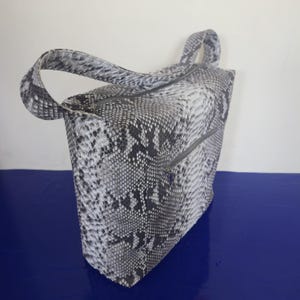 Może przedstawiać: Torba typu tote z wzorem wężowej skóry w odcieniach szarości i bieli. Torba ma dwa uchwyty i zapięcie na zamek błyskawiczny. Wykonana jest z teksturowanego materiału.