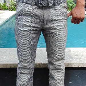 Op de afbeelding: Grijze en witte broek met slangenpatroon en bijpassende riem. De broek heeft een klassiek ontwerp met vijf zakken en een rechte pijp. De riem heeft een zilverkleurige gesp. De persoon draagt een zwart shirt.