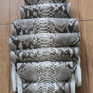 May include: Stacked rolls of python skin, showcasing a natural pattern of gray, black, and white scales. The rolls are arranged vertically, revealing the texture and design of the exotic leather. The background is a light brown wooden surface.