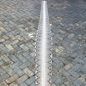 May include: A long, rectangular piece of python skin, with a pattern of black, grey, and white scales. The skin is laid out on a grey brick surface, showcasing its natural texture and colour variations. The snake skin is centered in the image.