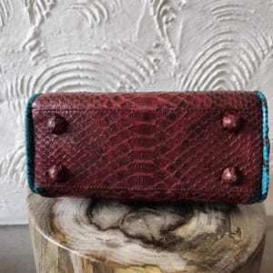 May include: A red snakeskin handbag with turquoise trim. The bag has a textured surface and four metal feet on the bottom.