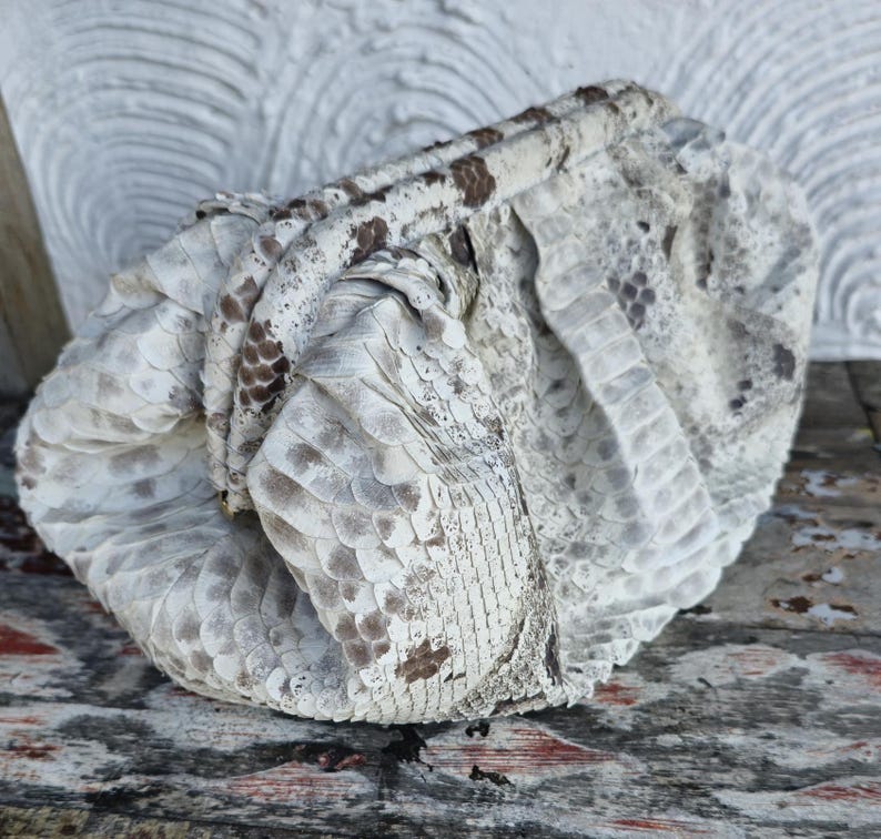 Może przedstawiać: Biała torebka ze sk&oacute;ry węża o fakturowanej powierzchni. Torebka jest złożona i ma brązowy pasek sk&oacute;rzany.