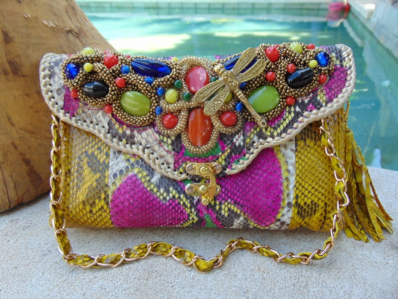 May include: A colourful clutch purse with a snakeskin pattern and a gold chain strap. The purse is decorated with a dragonfly charm and various colourful beads and stones.