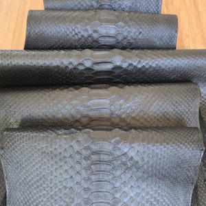 May include: A stack of black snakeskin leather pieces arranged in a cascading, overlapping pattern. Each piece displays a distinct, natural scale texture with a central ridge running vertically down the middle of the dark, matte-finished material.