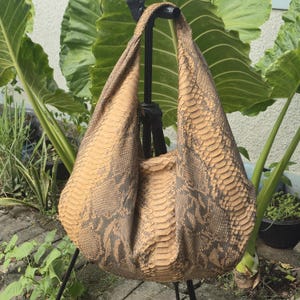 Puede incluir: Un bolso de hombro estilo hobo confeccionado en piel de serpiente texturizada en tonos beige, marrón claro y marrón oscuro. El bolso presenta una silueta holgada y desestructurada con un asa integrada y un patrón de escamas repetitivo.