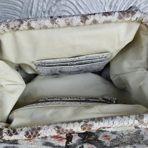 Może przedstawiać: Wnętrze białej torebki ze sk&oacute;ry węża z kieszenią zapinaną na zamek błyskawiczny i mniejszą kieszenią wewnętrzną. Torebka jest podszyta jasnobeżową tkaniną.