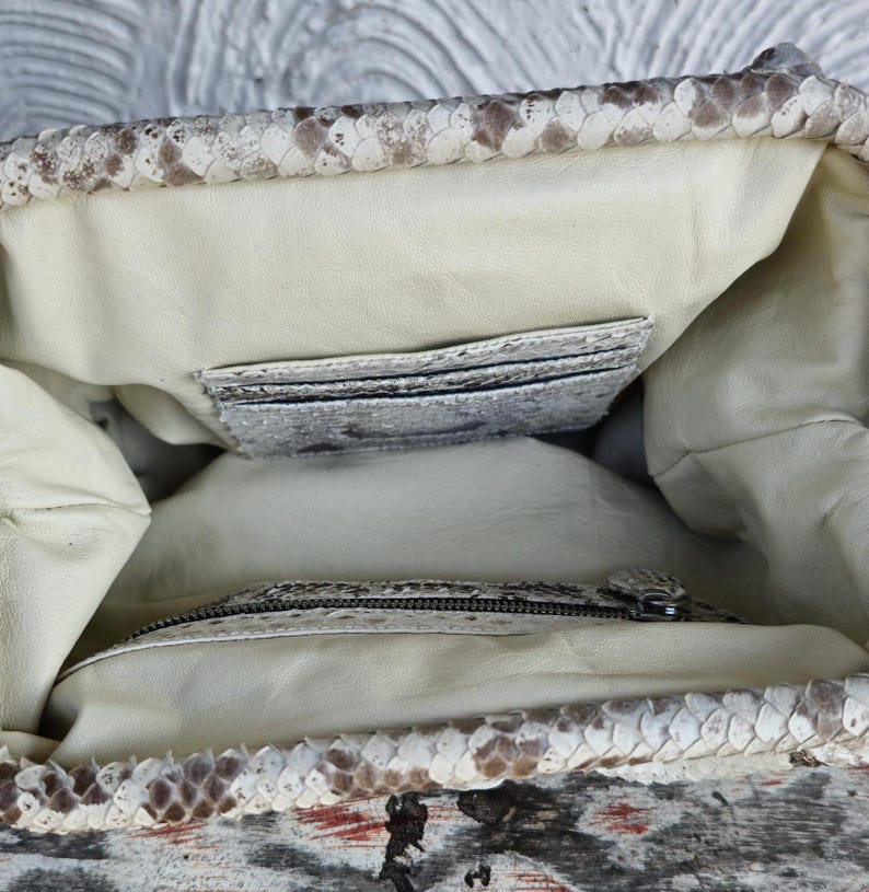 Może przedstawiać: Wnętrze białej torebki ze sk&oacute;ry węża z kieszenią zapinaną na zamek błyskawiczny i mniejszą kieszenią wewnętrzną. Torebka jest podszyta jasnobeżową tkaniną.