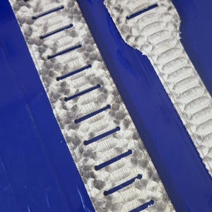 Könnte beinhalten: Zwei Gitarrengurte mit Schlangenhautmuster in Grau- und Weißtönen. Die Gurte haben mehrere vertikale Schlitze zur Anpassung und sind vor einem hellblauen Hintergrund platziert. Das Design ahmt die Textur von Reptilienhaut nach.