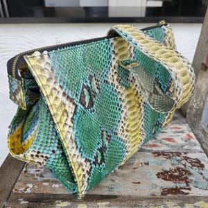 K&ouml;nnte beinhalten: Eine gr&uuml;ne, gelbe und braune Schlangenlederhandtasche mit einem Henkel oben. Die Tasche hat einen Rei&szlig;verschluss und ein einzigartiges geometrisches Design.