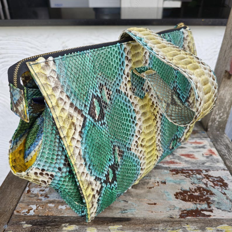 K&ouml;nnte beinhalten: Eine gr&uuml;ne, gelbe und braune Schlangenlederhandtasche mit einem Henkel oben. Die Tasche hat einen Rei&szlig;verschluss und ein einzigartiges geometrisches Design.