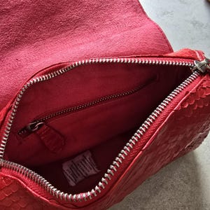 May include: Open view of a red leather handbag with a textured snakeskin pattern. The bag features a silver zipper closure and a matching red interior with a zippered pocket. The bag's flap is open, revealing the interior.