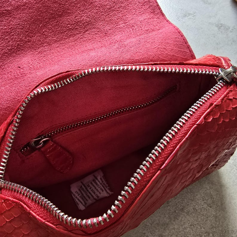 May include: Open view of a red leather handbag with a textured snakeskin pattern. The bag features a silver zipper closure and a matching red interior with a zippered pocket. The bag's flap is open, revealing the interior.