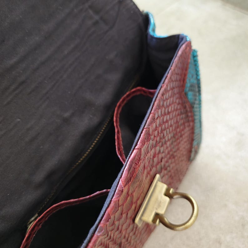 May include: A red, blue, and black snakeskin handbag with a gold clasp and a zipper closure. The bag has a black fabric lining and two interior pockets.