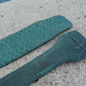 Könnte beinhalten: Zwei tealgrüne Lederriemen mit Schlangenhautmuster. Die Riemen liegen auf einer hellgrauen Oberfläche.