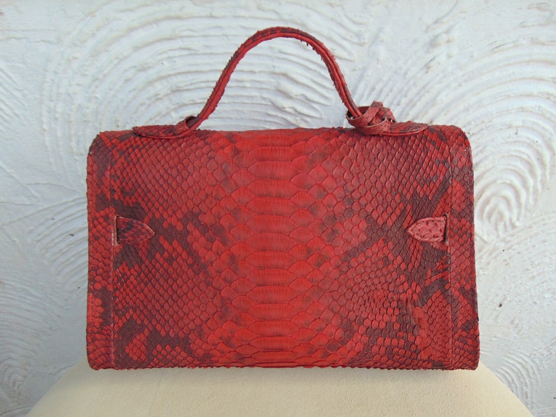 K&ouml;nnte beinhalten: Eine rote Schlangenlederhandtasche mit einem oberen Henkel. Die Tasche hat ein strukturiertes Schlangenledermuster und eine dunkelrote Farbe.