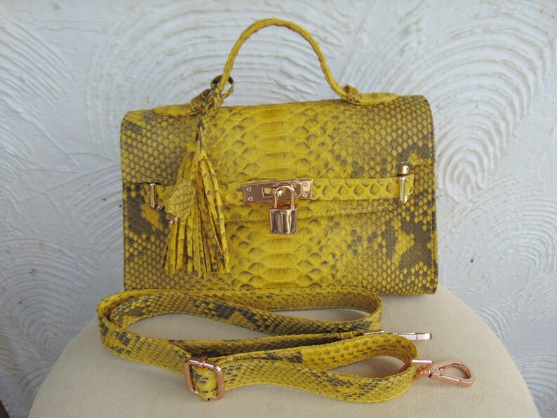 K&ouml;nnte beinhalten: Eine gelbe Schlangenleder-Handtasche mit einem goldenen Schloss und einem Quastenanh&auml;nger. Die Tasche hat einen Schulterriemen und einen Henkel.