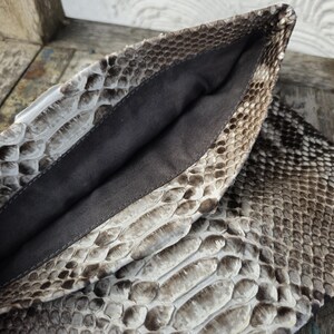 Pu&ograve; includere: Una pochette in pelle di serpente marrone chiaro con fodera in tessuto nero. La pochette &egrave; aperta e la fodera &egrave; visibile.