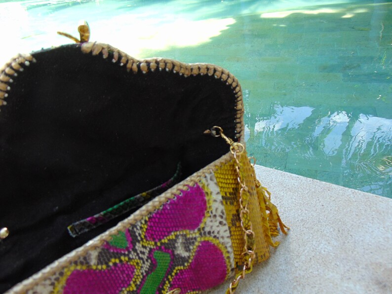 May include: A colourful clutch purse with a gold chain strap and a tassel. The purse is made from a variety of materials, including snakeskin, leather, and fabric. The purse is open and the inside is lined with black fabric.