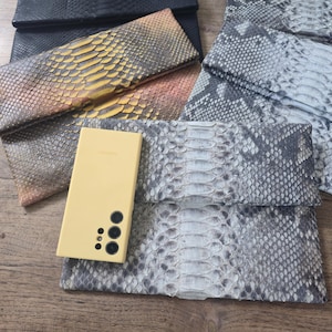 May include: A collection of rectangular clutch bags featuring textured snakeskin patterns in black, grey, and a gradient of yellow to brown. A pale yellow rectangular smartphone with a triple camera lens array is placed on top of the grey clutch.