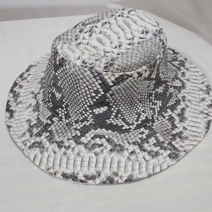 May include: A bucket hat with a snakeskin pattern in shades of grey and white. The hat has a wide brim and a rounded crown. The snakeskin design is detailed and textured, offering a stylish accessory.