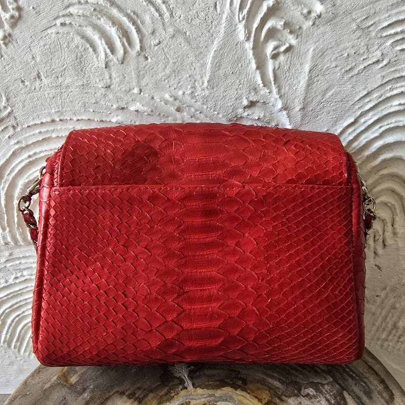 May include: A red handbag with a snakeskin pattern. The bag has a rectangular shape with a flap closure and a silver chain strap. The bag is made of a textured material and has a glossy finish.